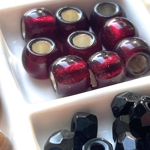 May include: Close-up of a white divided tray filled with various beads. The top section contains several dark red, round beads with a metallic core. The bottom section holds black, faceted beads. Other beads are visible.