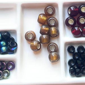 May include: A white divided tray filled with various beads. The beads are in shades of brown, black, purple, and iridescent blue. Some beads have a metallic finish, while others are translucent. The beads are round and cylindrical, suitable for jewelry making.