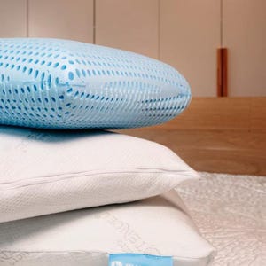May include: A stack of three pillows. The top pillow is light blue with a perforated, textured surface. The two bottom pillows are white with a smooth fabric cover, featuring a small blue tag with the white text BLU SLEEP on the bottom one.