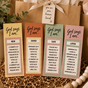 Puede incluir: Cuatro marcadores coloridos con el texto "Dios dice que yo soy..." en una escritura elegante. Cada marcador presenta un tema diferente: Nuevo, Salvado, Seguro y Amado, con versículos bíblicos correspondientes. Los marcadores se muestran con bolsas de regalo y acentos decorativos.