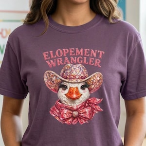Könnte beinhalten: Ein lila T-Shirt mit einer Grafik einer Gans mit Cowboyhut und Schal. Der Text "ELOPEMENT WRANGLER" steht über der Gans. Das Shirt ist aus weichem Material.