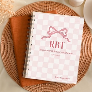Puede incluir: Un cuaderno de espiral con un patrón de cuadros rosas y blancos y el texto "RBT Registered Behavior Technician EST. 2025" se muestra en un mantel individual tejido. También se ven una taza de té, lápices y una piña.