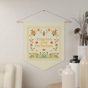 May include: A cream-colored banner with the words "Progress over Perfection" in yellow cross-stitch. The banner features bees and floral designs in yellow, orange, and green, hanging from a wooden rod.