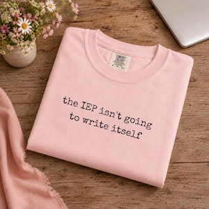 Puede incluir: Una camiseta de cuello redondo de color rosa claro con el texto impreso en negro 'the IEP isn't going to write itself' en la parte delantera. La camiseta tiene una etiqueta de la marca Comfort Colors en el cuello y está doblada sobre madera.