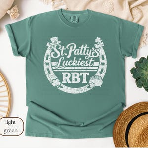 Puede incluir: Camiseta verde claro con un gráfico blanco que dice "St. Patty's Luckiest RBT" dentro de una herradura. El diseño incluye un sombrero de copa, tréboles y una olla de oro. La camiseta es de color verde claro liso.