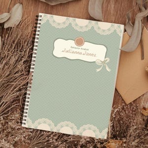 May include: A spiral-bound notebook with a pale blue cover and a lace design. The notebook has a label with the name "Julianna Jones" and the title "Behavior Analyst". The notebook is surrounded by dried flowers and leaves.