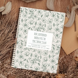 Könnte beinhalten: Ein Spiralnotizbuch mit einem floralen Muster in Grün- und Weißtönen. Das Notizbuch trägt den Text "REGISTERED BEHAVIOR TECHNICIAN" und "CHANGING LIVES ONE BEHAVIOR AT A TIME" auf einem weißen Etikett. Auf einer Holzoberfläche.