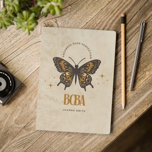 May include: A notebook with a butterfly design and the text "Progress Over Perfection" in a circular pattern. The butterfly is brown and gold. The notebook also has the text "BCBA" and "Joanna Smith". A pencil and pen are next to the notebook.