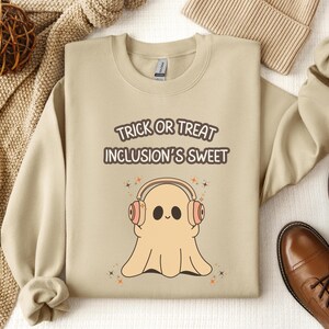 Puede incluir: Sudadera beige con el texto "Trick or Treat Inclusion's Sweet" y un fantasma de dibujos animados con auriculares. El fantasma tiene una cara sonriente y está rodeado de estrellas. La sudadera se combina con zapatos de cuero marrón y un gorro beige.