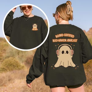 Puede incluir: Sudadera verde oscuro con un diseño de fantasma con auriculares y el texto "BOARD CERTIFIED BOO-HAVIOR ANALYST" en la espalda. Un diseño de fantasma más pequeño con "BCBA" está en la parte delantera. El diseño es en tonos beige, naranja y blanco.