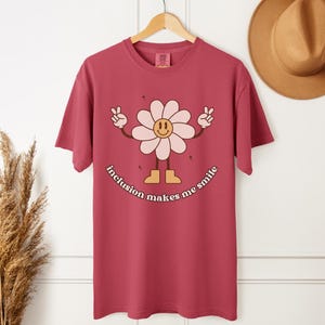 Op de afbeelding: Een bordeauxrood T-shirt met een lachend madeliefje dat vredestekens maakt. Het madeliefje heeft een lichtroze gezicht en bloemblaadjes, met de tekst "inclusion makes me smile" eronder.