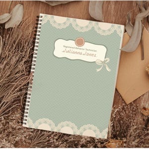 Puede incluir: Un cuaderno de espiral con una cubierta azul claro con diseño de encaje y el texto "Registered Behavior Technician" y "Juliana Jones." El cuaderno está rodeado de flores y hojas secas sobre una superficie de madera.