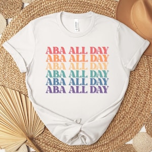 May include: White t-shirt with the text "ABA ALL DAY" in a rainbow of colors. The text is stacked vertically, with each line in a different color. The shirt is made of a soft material.