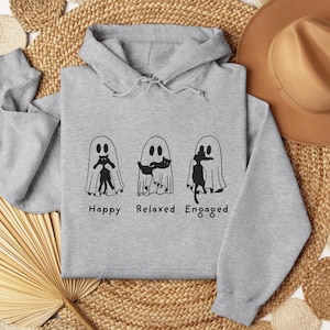 Puede incluir: Sudadera con capucha gris claro con un gráfico de tres figuras de fantasmas, cada una con un gato negro. El texto de abajo dice "Happy Relaxed Engaged". La sudadera está hecha de un material suave.