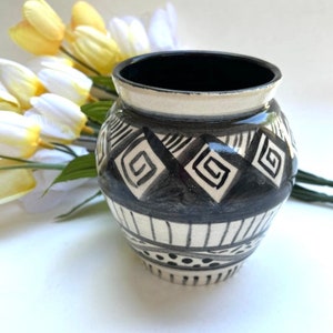 Pode incluir: Um pequeno vaso de cerâmica branca com um design geométrico preto e branco. O vaso tem um corpo redondo e uma boca larga.
