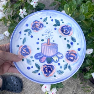 May include: A hand-painted ceramic plate with a light blue rim and a white center. The plate features a birthday cake with a lit candle, surrounded by blue and pink roses and green leaves. The plate is held by a person.
