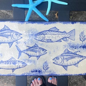 Puede incluir: Una bandeja de servicio rectangular de cerámica con base beige claro y un diseño azul pintado a mano que presenta cuatro ilustraciones de peces, bordes pintados con esponja azul y dos estrellas de mar azul brillante en el borde.