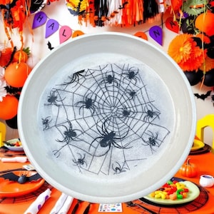 Pode incluir: Um prato redondo de cerâmica branca com uma estampa gráfica preta de uma teia de aranha detalhada com várias aranhas de tamanhos variados espalhadas pela superfície. O prato tem uma borda larga e lisa e um design circular limpo.
