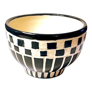May include: A ceramic bowl featuring a black and white checkered pattern around the upper rim and a series of vertical black and white stripes on the lower portion. The interior of the bowl is a solid off-white color with a smooth, glossy finish.