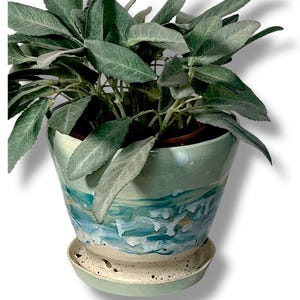 Peut inclure: Une plante en pot avec de grandes feuilles vertes texturées. Le pot en céramique est peint avec un motif marin dans des tons de bleu, blanc et vert. Une soucoupe assortie est placée en dessous.