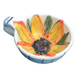 May include: A white ceramic bowl with a blue and white striped rim. The bowl has a handle and features a hand-painted sunflower design with yellow petals, orange accents, and a brown centre.