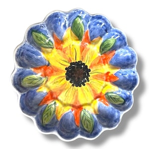 Op de afbeelding: Een keramische plaat met een zonnebloemontwerp. Het midden is geel met een zwart centrum, omgeven door oranje bloemblaadjes. De buitenrand is blauw met groene bladaccenten. De plaat heeft een geschulpte rand.