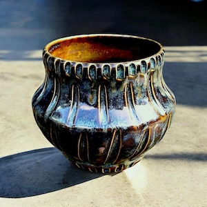 Puede incluir: Una maceta de cerámica azul oscuro y marrón con un diseño texturizado y acanalado. La maceta tiene una boca ancha y una base redondeada.