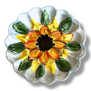 Könnte beinhalten: Ein handbemalter Keramik-Servierteller mit Sonnenblumen-Design. Der Teller hat einen gewellten Rand und eine zentrale Sonnenblume mit gelben, orangefarbenen und braunen Blütenblättern, umgeben von grünen Blättern. Der Hintergrund ist hellgrau.