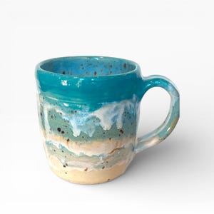 Può includere: Una tazza in ceramica fatta a mano con bordo e manico blu turchese. La tazza presenta un design costiero con sfumature di blu, bianco e beige, che ricorda le onde dell'oceano. La tazza è alta circa 10 cm e ha un manico curvo.