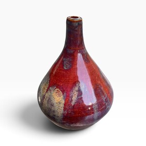 May include: A teardrop-shaped ceramic vase with a glossy finish, featuring a deep red and burgundy glaze that transitions into mottled patches of cream and dark brown near the base, with a narrow neck and a small circular opening at the top.