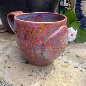 Puede incluir: Una taza de cerámica hecha a mano con un diseño único. La taza presenta un esmalte marrón rojizo con detalles azules y naranjas. El exterior está decorado con líneas talladas y un motivo floral. La taza tiene un asa grande.