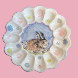 Op de afbeelding: Een witte keramische eierenschaal met een geschulpte rand en een geschilderd konijn in het midden. De schaal heeft 12 eierinkepingen en is versierd met pastelkleurige stippen.