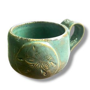 Puede incluir: Taza de cerámica hecha a mano con un esmalte verde agua. La taza presenta un diseño de dólar de arena en relieve y un asa redondeada. El borde y los detalles del dólar de arena tienen un acento marrón dorado. Adecuada para bebidas frías o calientes.