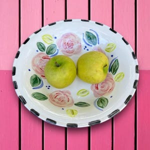 May include: A white ceramic serving plate with a black and white checkered rim, decorated with pink roses, green leaves, and two green apples. The plate is set against a pink wooden background.