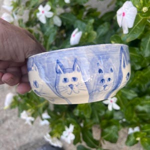 May include: A ceramic bowl with a light blue rim and a cream-colored interior. The bowl is decorated with blue cat faces around the exterior. The bowl is held by a person's hand, with green foliage and white flowers in the background.