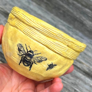 May include: A yellow bowl with a textured, ridged rim and a smooth, rounded body. The exterior features two black ink-style illustrations of bees, one large and one small, positioned on the side of the bowl against the bright yellow surface.