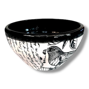 May include: A ceramic bowl with a glossy black interior and rim. The exterior features a white base with intricate black ink illustrations of a bird, stylized leaves, and cursive handwritten text wrapping around the curved surface of the bowl.