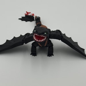 May include: A black dragon figurine with a wide, toothy grin and green eyes. The dragon has large, spread wings and a red and white flame detail on its tail. It is posed on a white surface.