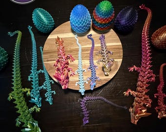 May include: A collection of colorful, articulated dragon figurines and dragon egg sculptures. The dragons are in various colors, including green, blue, red, and purple. The eggs are blue, green, and rainbow-colored. The image includes the text "AUTHORIZED SELLER".