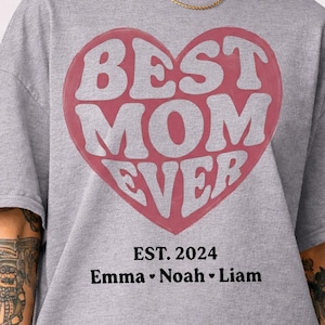 Puede incluir: Camiseta gris con un gran diseño de corazón rojo que dice "BEST MOM EVER" en letras blancas. Debajo del corazón, el texto dice "EST. 2024" y los nombres "Emma Noah Liam".