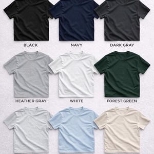 Puede incluir: Exhibici&oacute;n de nueve camisetas de manga corta en varios colores: negro, azul marino, gris oscuro, gris jaspeado, blanco, verde bosque, ceniza, azul claro y arena. El texto "AVAILABLE COLORS" est&aacute; en la parte superior.