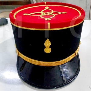 以下が含まれることがあります： 金色のトリムとディテールが施された、黒と赤のミリタリースタイルの帽子。帽子は、光沢のある黒いブリム、金色の刺繍が施された赤いトップ、前面に金色のエンブレムが特徴です。フォーマルなアクセサリーです。
