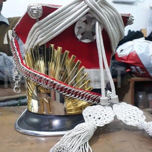 Puede incluir: Un sombrero militar ceremonial con una corona de terciopelo rojo, adornos plateados y una visera dorada. El sombrero presenta una pluma blanca, cordones plateados y detalles metálicos decorativos. Una cadena y una borla cuelgan del sombrero.