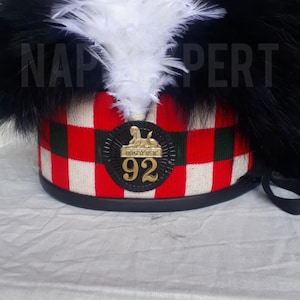 May include: A military hat featuring a red and white chequered design, black fur trim, and white feathers. A gold emblem with the number 92 is centered on the front. The text "NAP PERT" is visible on the hat.