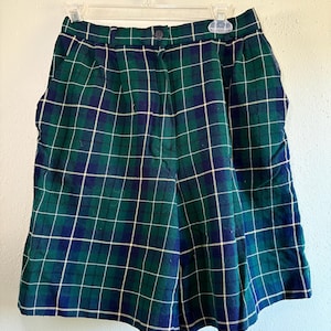 May include: Green, navy, and white plaid shorts hanging on a clear plastic hanger. The shorts have a button closure and pockets. The plaid pattern is a classic design, suitable for a variety of outfits.