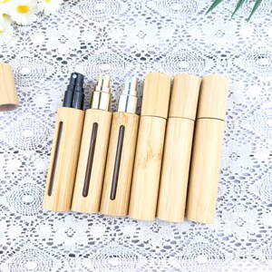 May include: A collection of bamboo travel-size perfume atomizers. The atomizers have a natural wood finish with a vertical viewing window. The spray nozzles are black, gold, and silver. The atomizers are displayed on a white lace surface.