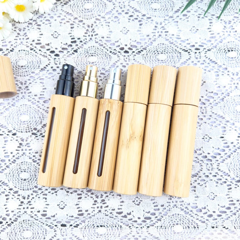 May include: A collection of bamboo travel-size perfume atomizers. The atomizers have a natural wood finish with a vertical viewing window. The spray nozzles are black, gold, and silver. The atomizers are displayed on a white lace surface.