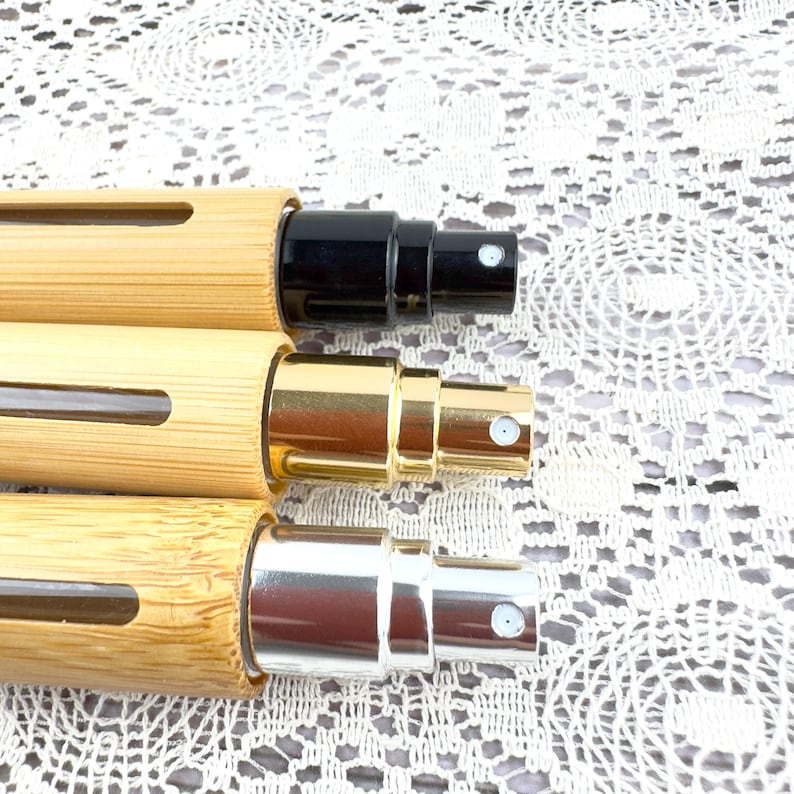 May include: Close-up of three perfume atomizers. The atomizers have bamboo-style casings with a rectangular window. The spray nozzles are black, gold, and silver. The background is a white lace pattern.