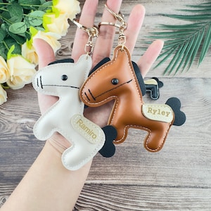 May include: Two leather horse keyrings, one white and one brown, with gold-coloured key rings. The white keyring has the name "Ramiro" and the brown keyring has the name "Ryley". Both have black manes and tails.
