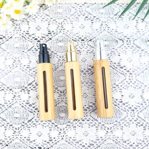 May include: Three bamboo spray bottles with different colored tops: black, gold, and silver. Each bottle has a rectangular window showing the liquid level. The bottles are arranged on a white lace surface.